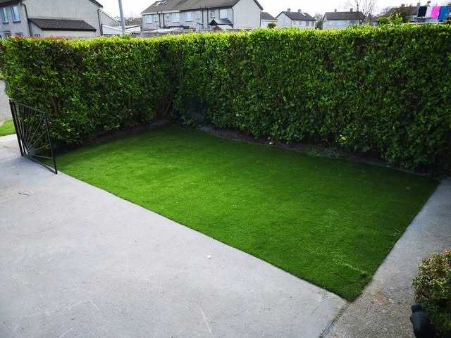 Artificial grass laid over a concrete yard beside a tall green hedge