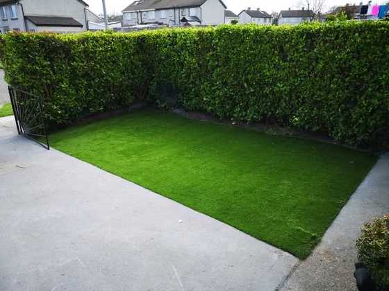 Artificial grass laid over a concrete yard beside a tall green hedge